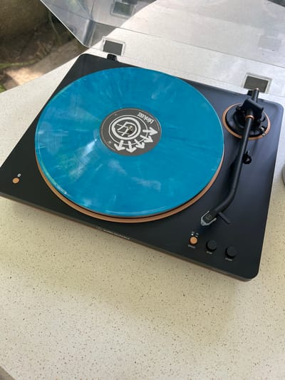 The image shows a black and brown Audio-Technica AT-LP7 turntable with a clear dust cover in the upper left. A blue vinyl record with a white label featuring a logo with arrows (likely Blink-182's Nine album) is on the turntable. The turntable is on a speckled countertop.
The image does not contain people, actions, or interactions.
The location is impossible to assess from the provided image.