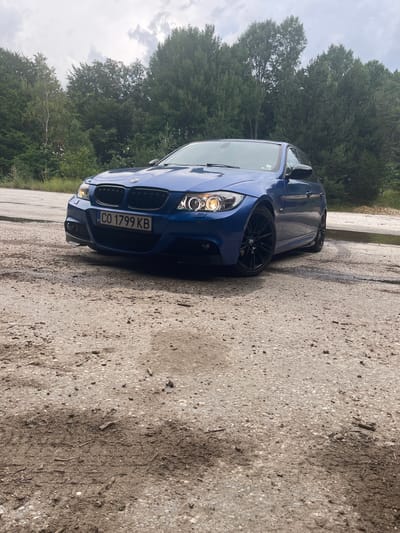 The image shows a blue BMW M3 parked on a gravelly surface next to a paved area. The car is facing towards the left of the frame, with its headlights on. It has black wheels and a license plate that reads "CO 1799 KB". In the background, there are dense green trees and a cloudy sky. The scene appears to be outdoors, possibly in a rural or semi-rural setting in Panagyurski kolonii, Bulgaria, given the foliage and the type of road surface. The lighting suggests it could be daytime, perhaps overcast due to the clouds. There are no people visible in the image, and the focus is solely on the car and its surroundings. The overall impression is a static shot of a parked car.