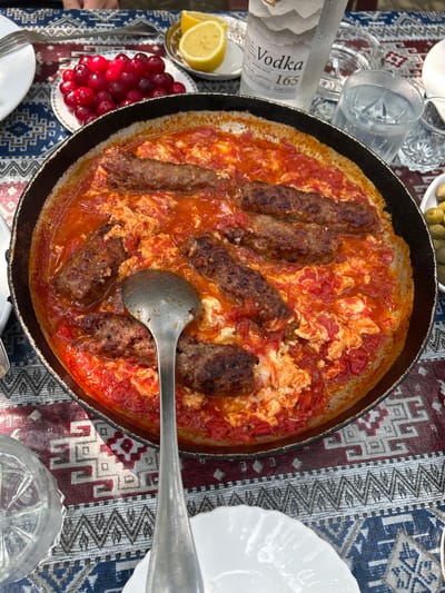 This image shows a close-up of a meal served in a traditional Azerbaijani setting. The main subject is a rustic, dark-colored skillet filled with a dish that appears to be kuku seba, a type of Persian frittata, or perhaps a similar egg and meat dish cooked in tomato sauce. Several elongated, browned meatballs or kofta are nestled within the mixture of scrambled eggs and a vibrant red tomato sauce, which has a slightly chunky texture. A large, metallic ladle rests in the skillet, suggesting the dish is ready to be served or is in the process of being eaten.

Surrounding the skillet, the setting is revealed to be a table covered with a richly patterned tablecloth featuring geometric designs in red, white, and blue. To the left of the skillet, a bottle of "Vodka 165" is visible, along with a partially sliced lemon and a small white dish filled with bright red cherries or cranberries. Several glasses, one containing ice and water, are also scattered around the table. In the upper right corner, a small white bowl holds green olives.

The lighting suggests it might be daytime, possibly outdoors given the dappled sunlight that creates shadows on the tablecloth. The overall impression is of a casual, shared meal, possibly a family gathering or a meal with friends, set in a warm and inviting atmosphere. The presence of traditional elements like the patterned tablecloth and the food itself hints at a cultural context, which is further supported by the provided location context of Sabail Raion, Azerbaijan. No specific emotions are conveyed, but the presentation suggests a hearty and appealing meal.