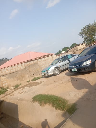 This image captures an outdoor scene in Gwagwa, Nigeria, likely during the day. Two cars are parked on a dirt lot in front of a building with a prominent red corrugated metal roof and a stone block wall. The car in the foreground is a dark gray sedan, possibly a Honda Accord, with a license plate that reads "ABC 365 JZ". The car behind it is a silver sedan, appearing older and slightly weathered, with a turquoise accent on its front bumper.

The ground is a mixture of dirt and some patches of dry grass, with a concrete drainage channel running along the bottom of the frame. In the background, there are more buildings and a large tree. The sky is a clear, pale blue with a few wispy clouds, suggesting fair weather. The lighting indicates it is daytime, possibly late morning or early afternoon, with shadows cast by the cars and an unseen object, possibly the photographer. No people are visible in the image. The overall impression is one of a simple, everyday scene in a residential or commercial area.