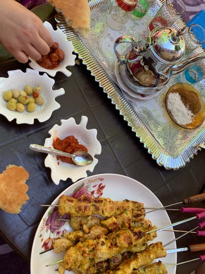 A table laden with food suggests a meal taking place in Sidi Slimane Moul Al Kifane, Morocco. A silver tray holds a teapot, glasses, and a dish of what appears to be salt and sugar. A plate of skewers sits near dishes containing olives and a red paste, possibly a condiment. A hand reaches for the food, and bread is also present.