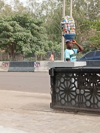 The image depicts an urban street scene in Darmanawa, Nigeria, under bright daylight. In the foreground, on the right, a black metal gate with intricate hexagonal and star-like geometric patterns is partially visible, secured with a silver padlock. The ground consists of concrete paving in the immediate foreground, transitioning to a paved asphalt road surface.
The central subject is a young male, dark-skinned, positioned in the mid-ground, facing towards the right. He wears a light blue t-shirt with a green and white horizontal stripe design and dark trousers. He is balancing a substantial, tall stack of various packaged consumer goods, including what appears to be snacks, instant noodles, or beverages, contained within and piled above a blue woven basket on his head. His left hand is raised, lightly touching his forehead, seemingly for balance or adjustment of the load.
Behind him, across a concrete median barrier, another individual is visible in the mid-background. This person, dressed in light orange or brown attire, is seated or crouching on the far side of the barrier, facing away from the camera. The median barrier has political posters affixed to it, displaying multiple male faces. A dark blue sedan is partially visible behind the median, on the far side of the road. A utility pole stands vertically in the background, near the far side of the road.
The background is dominated by dense green foliage and numerous trees, suggesting a vegetated roadside or park area. The scene conveys a moment of daily commerce in a public thoroughfare.