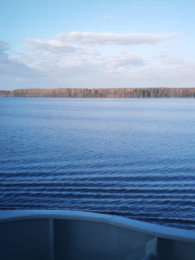 The image presents a serene, expansive landscape featuring a wide body of water, a distant treeline, and a partly cloudy sky, viewed from the deck of a boat. The scene is set in what appears to be a natural, undeveloped area, with the location context being Andrukovo, Russia.
The upper half of the frame is dominated by a light blue sky, adorned with scattered white and soft grey clouds that stretch horizontally across the expanse. Below the sky, a dense, dark treeline forms a continuous band along the distant horizon, marking the edge of the land. The trees appear to be predominantly deciduous, many with bare branches, suggesting a season of late autumn or early spring, though some darker evergreens might be interspersed.
The vast body of water, likely a wide river or a large lake, occupies the mid-ground and much of the foreground. Its surface is a deep, tranquil blue, characterized by numerous small, parallel ripples and waves that add texture and movement to the scene. In the immediate foreground, at the bottom of the image, the grey-painted metal structure of a boat is visible, including a section of its railing and inner hull, from which the photograph was taken.
No people or specific activities are depicted, conveying a sense of calm and quiet observation. The lighting suggests it is daytime, possibly late morning or early afternoon, under fair weather conditions. There is no visible text present in the image.