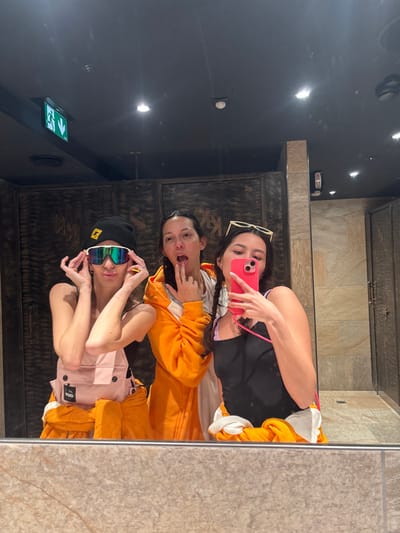 Three women take a selfie in a mirrored room. The woman on top holds a pink phone. The woman in the middle is wearing an orange outfit and sticking her tongue out. The woman on the bottom is wearing a pink and yellow outfit with sunglasses and a black beanie. The wall behind them is a dark color with an exit sign and round lights. The room also has tan tiling.