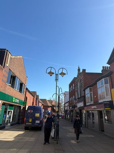 The image displays a daytime street view in Crewe, United Kingdom, under a clear blue sky. The scene is a commercial street lined with multi-story brick buildings. On the left, a building houses a Lloyds bank branch with its prominent green and white signage. Adjacent to the bank, a dark blue Mercedes-Benz Sprinter van, marked with "G4S" branding and registration LM19 AJO, is parked on the pavement, facing away from the camera.
Several ornate street lamps with twin, upward-curved lanterns stand along the center of the street. In the immediate foreground on the left, a male individual, dressed in a dark t-shirt, dark trousers, and sunglasses, stands on the pavement. He holds a white bag in his left hand and a dark mobile phone to his right ear. On the right side of the foreground, another male individual, wearing a dark long-sleeved top and dark trousers, walks away from the camera.
The right side of the street features various shopfronts, including an "ADMIRAL SLOTS" casino with a black and yellow sign, situated below a building with a first-floor bay window. Further down the street, additional pedestrians are visible. Sunlight casts distinct shadows across the light-colored flagstone pavement.