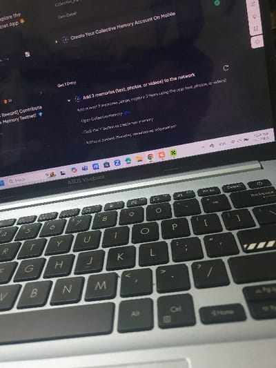 The image shows a laptop computer with a black keyboard and silver chassis. The screen displays text related to a website called "Collective Memory," with instructions on how to create an account and add memories to the network. The laptop is open and the keyboard is visible, with the keys "O," "P," "[," "]", "?", and "/" being in focus. The location is an unspecified indoor space within the district of Kecamatan Jombang in Indonesia.  The coordinates provided (-7.519 latitude, 112.236 longitude) are indicative of the location but do not provide a specific address.  The environment is a typical indoor workspace setting, possibly a home or an office.
