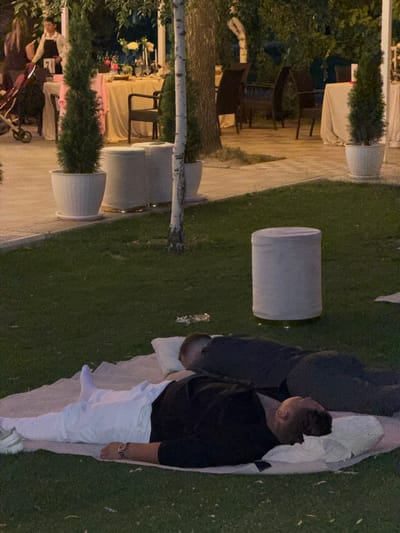 In the city of Kara-Jyrgach, Kyrgyzstan, an outdoor event is taking place in what appears to be a garden or park setting. In the foreground, two individuals are lying down on a picnic blanket on a grassy area. One person, dressed in white pants and a dark shirt, is lying on their back with their head resting on a white pillow. They are wearing a watch on their left wrist. The other person, also in dark clothing, is lying next to them. In the background, there are trees, potted plants, and a table set for a meal. Several chairs are visible around the table, and a person in an apron is standing near it. The overall atmosphere suggests a relaxed gathering or a catered event.