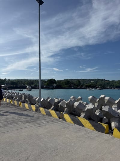 This image depicts a pier or dockside area in Toyapakeh, Indonesia, under clear daylight conditions. The foreground consists of a grey concrete pier surface with visible textures and shadows. Along the right edge of the pier, a series of interlocking, multi-faceted grey concrete tetrapods form a barrier or breakwater. These tetrapods feature diagonal yellow and black striped paint at their base. Approximately in the center of the image, a tall, slender metallic pole, likely a lamppost, extends vertically from the pier towards the sky, topped with a dark, rectangular fixture.
To the far left, a partial view of an individual, dressed in dark clothing, is visible, walking away from the camera. Beyond the pier and tetrapods lies a body of turquoise-blue water, indicative of a strait or bay. Several boats are present on the water; a white motorboat with a cabin is visible in the mid-distance, moving from left to right. Other smaller vessels are moored or scattered closer to the distant shoreline. The background features a lush, green, and hilly coastline covered with dense vegetation and trees. The sky above is a bright blue with scattered, wispy white clouds. The overall scene suggests a coastal port or transit point.