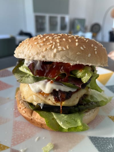The image displays a single, fully assembled hamburger resting on a geometrically patterned plate, in an indoor setting located in Foetz, Luxembourg. The hamburger is constructed from a sesame seed bun, with the top bun visible. Layered within are green and dark red lettuce leaves, a generous application of red sauce (appearing to be ketchup), and a creamy white sauce (consistent with mayonnaise or aioli). Below these, a thick, light-colored patty, possibly chicken or halloumi, is covered with melted yellow cheese. A dark, viscous, amber-colored sauce drips from the side of the patty. Underneath the patty are dark, round slices, indicative of grilled vegetables such as eggplant, followed by more green lettuce and the bottom bun. The plate features a repeating pattern of pastel pink, yellow, and grey triangles. The background is severely out of focus, showing indistinct light-colored walls and blurred structural elements or furniture, suggesting a contemporary interior. No individuals are present within the frame.