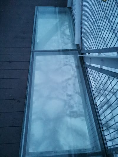 The image displays an overhead, downward-angled view of a section of an outdoor glass-bottomed walkway or bridge, situated in Мастоўскі сельскі Савет, Belarus. The left side of the frame features a dark, horizontally planked wooden surface. Adjacent to this, in the center, are two elongated, rectangular glass panels arranged in sequence, forming a transparent segment of the walkway. The glass exhibits a degree of translucence and surface irregularities, through which a textured, white, and uneven landscape, consistent with snow or ice-covered ground, is visible below. To the right of the glass panels, a light-colored metal railing with a diagonal grid pattern extends, featuring vertical support posts. The overall environment is an outdoor winter scene under diffused, cool-toned lighting. No individuals, specific actions, or interactions are present in the frame.