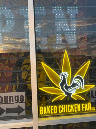 The image shows the exterior of a business in Sunland Park, New Mexico, USA. A neon sign in yellow and white depicts a cannabis leaf with a rooster silhouette in the center, and the text "BAKED CHICKEN FAR..." below it. Behind the neon sign, a large graphic of a face, possibly of Bob Marley, is visible. To the left, a sign reads "lounge" with an arrow pointing to the right. The business appears to be located on a street, as indicated by the reflection of the sky in the window. The overall impression is that of a quirky, potentially cannabis-themed establishment.