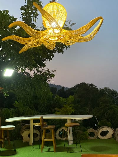 The image shows an outdoor scene in Khlong Khian, Thailand at dusk. Dominating the left side is a large, illuminated, yellow-wicker sculpture resembling a crab. It appears to be mounted on or near a tree, with the sky visible behind it. The right side of the image features a seating area with white tables and wooden stools, some arranged around circular tree-stump elements. Lush greenery fills the background, obscuring much of the landscape. A light source is visible near the bottom center of the image, illuminating the surrounding foliage.