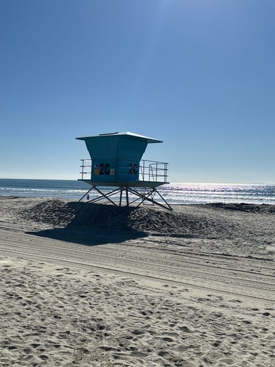 A light blue lifeguard tower stands on a sandy beach next to the ocean. The tower has the number 26 displayed on its side. The sand shows tire tracks, and the ocean reflects the sunlight with a shimmering effect. The sky is clear blue.