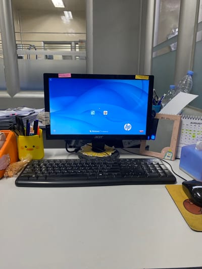The image displays an unoccupied office workstation, viewed from a direct, frontal perspective. The specific location within the city of Cipeundeuy, Indonesia, cannot be determined from the visual evidence.
The central focus is a black Acer computer monitor displaying a Windows 7 Professional desktop with a blue wave background and an HP logo. A small pink sticky note and a small yellow sticky note are adhered to the top bezel of the monitor. A black Logitech full-sized keyboard is centered on the light-grey desk surface directly in front of the monitor. Beneath the monitor's stand, a circular black coaster with a yellow sunflower design is visible.
To the left of the keyboard, an orange plastic container holds writing implements, positioned next to a yellow, duck-shaped pen holder also containing pens. A clear plastic bag filled with light-colored, granular snack items rests near the duck pen holder. Behind these items, a stack of papers and folders is visible.
To the right of the keyboard, a black computer mouse is positioned on a yellow mousepad adorned with a brown character graphic. Behind the mousepad, a small, upright, rectangular mirror with a light-pink frame and a penguin illustration is present. A white spiral-bound calendar, displaying "AUGUST" at its top, stands behind the mirror, adjacent to a light-blue tissue box. Two clear plastic water bottles are partially visible behind the calendar and tissue box.
The background indicates an office environment, featuring light-colored partition walls, some of which include frosted or translucent glass panels. Overhead fluorescent lighting fixtures are reflected in these panels, and a glimpse of another office area is visible through the glass. No individuals are present in the scene.