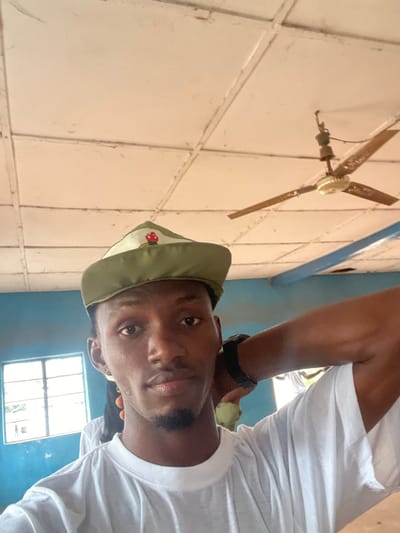 A young Black man wearing a green cap and a white t-shirt takes a selfie indoors. The man has short black hair, brown eyes, and a goatee. He is looking directly at the camera with a neutral expression. His left arm is bent behind his head, and he is wearing a black watch on his left wrist. In the background, there is a white ceiling with a fan, a blue wall, and a window with bars. The scene appears to be a classroom or a simple room.