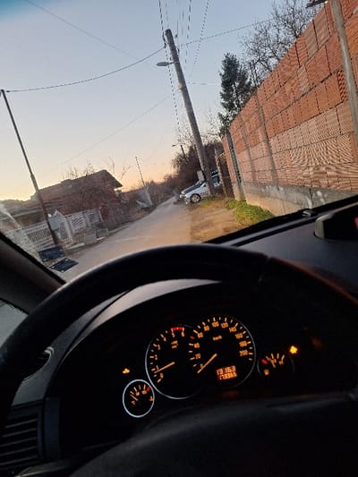 The image presents a static scene captured from the interior of a stationary vehicle, looking forward onto a residential street in the city of Katina, Bulgaria, during twilight. The sky overhead transitions from pale blue to an orange-pink hue near the horizon.

In the foreground, the upper portion of a black, three-spoke steering wheel is visible on the left. The vehicle's instrument cluster, centrally positioned, is illuminated with orange backlighting. The speedometer displays 0 km/h, and the tachometer indicates 0 RPM. A digital display within the cluster shows "13:16" (time) and "173365" (odometer reading). The fuel gauge indicates approximately a quarter-tank, and the coolant temperature gauge shows a reading slightly above its lowest mark. Three yellow warning lights are illuminated: one resembling a vehicle with an arrow (possibly ESP or traction control), a general warning light, and a wrench or engine symbol.

The exterior view shows a narrow road, likely unpaved or poorly maintained, receding into the distance. On the left side of the road, multiple residential houses with various roof types are visible, interspersed with fences and bare deciduous trees. A utility pole with overhead wires stands on the left. On the right side of the road, a tall, unfinished wall constructed from stacked red hollow bricks rests upon a concrete foundation. Behind this wall, an evergreen tree and additional bare trees are present. A wooden utility pole, equipped with a street light and multiple power lines, stands on the right, leaning slightly. Further down the street on the right, two parked vehicles are identifiable: a light-colored sedan with distinct yellow or gold aftermarket wheels, and a second, darker vehicle partially obscured behind it.
