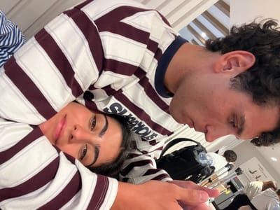 This image captures a close-up, overhead view of a young couple, seemingly on vacation or traveling. The woman is positioned lower in the frame, her face turned upwards and partially obscured by the man's arm and a striped garment. She has dark hair and is wearing a striped shirt with maroon and white stripes, which also drapes over her. Her eyes are open and she appears to be looking towards the camera.
The man is positioned above and to the right of the woman, his head and shoulder dominating the upper right portion of the image. He has dark, curly hair and is also wearing a similar maroon and white striped shirt, with a dark blue collar. His face is in profile, looking downwards, and his arm is extended, resting on or near the woman.
In the background, hints of an indoor environment are visible. There are luggage items, including a black backpack with a water bottle. Further back, other people and what appear to be airport or terminal-like structures are present, including signage with English text, one of which reads "London. Corn & Bread." The lighting suggests an indoor, possibly public space, illuminated by overhead lights. The overall impression is an intimate moment captured during travel.
While the image context suggests a travel scenario, there is no visual information within the image that specifically indicates the location as Mykonos, Greece.
