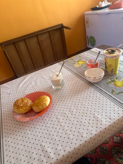 A table is set with a white and gray polka-dot tablecloth. A container of food, a glass of pink liquid with a spoon, and a white bowl are in the top left corner. Further down is a glass filled with sugar and a spoon, next to a plate with two buns, one with sesame seeds. A wooden frame is attached to the wall on the left side of the image.
