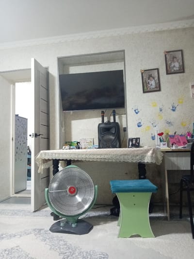 This image depicts an interior room in Shirin, Uzbekistan. The scene is presented from a low-angle perspective, facing towards the back wall. The room features light-colored, possibly textured wallpaper with a white decorative molding along the ceiling line. The wall on the right displays multiple blue and yellow handprint decorations, with two framed photographs of individuals hung above them.
In the center, a dark-legged table, covered by a light-colored, lace-edged tablecloth, is positioned. Above the table, a black flat-screen television is mounted within a recessed opening in the wall. On the tablecloth, directly beneath the television, rests a black portable speaker accompanied by two microphones. Various small objects, including what appears to be books or papers, are scattered on the tablecloth, along with a desk organizer containing writing implements and a red artificial flower or plant on the far right section of the table.
On the floor in the foreground, a mint green retro-style floor fan with a chrome-colored protective grill and a red central logo is prominently placed on a light-colored carpet with a subtle patterned design. To the right of the fan, a light green, hourglass-shaped stool with a blue upholstered seat is positioned. A dark, metal-framed chair is partially visible behind the stool on the far right.
An open, light-colored interior door with horizontal decorative panels and a dark handle is visible on the left, leading into another brightly lit room. A partially visible white appliance, resembling a refrigerator or tall cupboard with items on its surface, is visible in the room beyond the door. No people, actions, or direct interactions are visible in the scene.