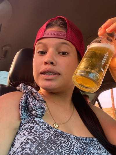 A close-up, sideways-oriented photograph captures a young woman inside a car. She is holding a clear glass bottle containing a golden, frothy beverage, likely beer, with her hand positioned above her head. The woman has dark hair, a light complexion, and is wearing a red mesh baseball cap and a patterned top featuring a dark and light speckled design. A gold necklace with a circular pendant is visible around her neck. She presents a relaxed expression with a slight smile, revealing braces on her teeth.

The setting appears to be the interior of a vehicle, with a dark headrest visible in the background. Bright natural lighting suggests the photo was taken during the day.

Text on the bottle, identified through OCR, includes "CERVEZA DE CALORÍA REDUCTA" (Reduced Calorie Beer), "CERVEZA PILSEN (100) NORMAL: CALORIAS" (Pilsen Beer (100) Normal: Calories), and "BAJA EN CALORÍAS: 29 CALORIAS" (Low in Calories: 29 Calories), indicating a low-calorie pilsen beer. The partial phrase "VIVEY DISPO" is also present on the label. This casual moment is situated in La Cruz del Pastel, Venezuela.