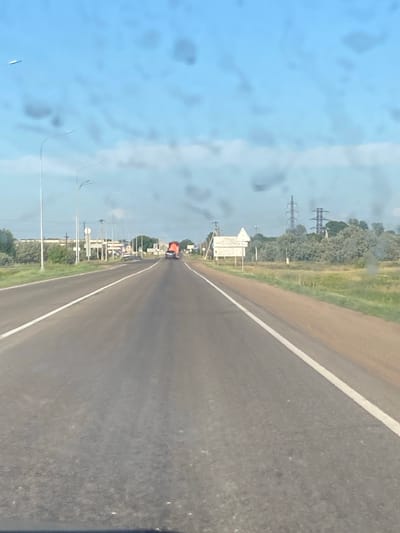 The image depicts a view from a vehicle traveling on a paved road. The road has a white line dividing the lanes, and the surrounding environment consists of green vegetation and trees. In the distance, power lines and transmission towers are visible, suggesting an open, rural or semi-rural setting. There are also streetlights lining the road.
A bright orange vehicle is seen ahead on the road, along with other vehicles further in the distance. The sky is blue with some clouds, indicating it is daytime. The perspective suggests the photograph was taken from the passenger seat of a car, looking forward and slightly to the left. The image has a slightly blurred or smudged quality, possibly due to dirt or water droplets on the windshield.
The scene is likely located in Rubtsovsk, Russia, given the provided location context. No people are clearly identifiable in the image, but the presence of vehicles and infrastructure suggests human activity. The weather appears to be fair. No prominent text is legible in the image.