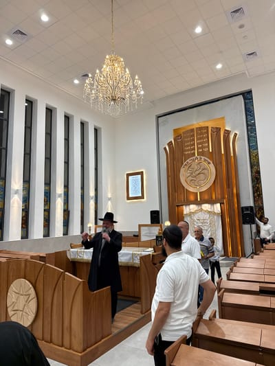 This image depicts the interior of a modern synagogue located in Beer Yaaqov, Israel. The scene features a spacious, brightly lit hall with high white walls and a white grid ceiling fitted with recessed lighting. A large, multi-tiered crystal chandelier hangs prominently from the ceiling in the center.

Along the left wall, several tall, narrow windows with vertical stained glass panels are visible, with wall-mounted sconces positioned between them. The rear wall is dominated by a grand, elaborately carved wooden Aron Kodesh (Holy Ark), featuring vertical slats, carved Hebrew text (likely the Ten Commandments), and a central circular motif of a stylized lion. Dark stained glass panels flank the Aron Kodesh. To its left, an illuminated framed Hebrew text is mounted on the wall. Black audio speakers on stands are positioned near the Aron Kodesh.

In the mid-ground, on an elevated wooden Bima (speaker's platform) draped with a white and gold cloth, a man, presumably a Rabbi, is actively speaking. He is bearded, wears a black fedora, a black long coat, and holds a microphone in his left hand while gesturing with his right.

Several individuals are present in the foreground and mid-ground, primarily men. One man with a dark beard and a white polo shirt stands with his back to the camera in the immediate foreground, facing the speaker. Other men, mostly in casual attire such as polo shirts, and some wearing yarmulkes, are visible standing in the aisles to the right, observing the speaker. A young boy is also visible among the adults in the mid-ground. Rows of light-colored wooden pews with individual seating are arranged on the right side of the frame.