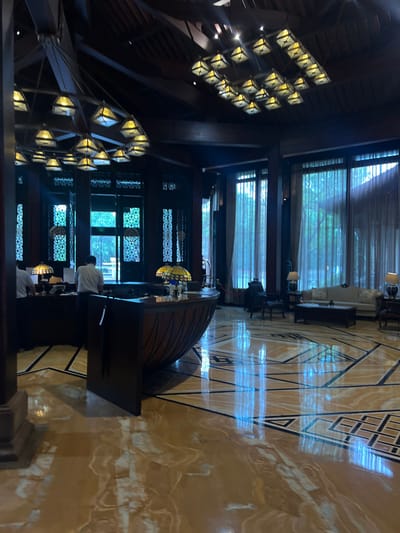 This image displays the interior of a grand lobby or reception area, located in Tongxiang, China. The scene is dominated by a high, dark timber ceiling featuring exposed beams, from which multiple large, multi-lamp chandeliers are suspended. Each chandelier consists of numerous geometric, square-based pyramid light fixtures.

The floor is composed of highly polished light brown or golden marble, inlaid with dark geometric patterns and lines, creating significant reflections of overhead and window light. In the foreground, a prominent, dark wooden reception desk with a curved, ribbed front is positioned centrally. Two Tiffany-style lamps with domed, stained-glass shades sit atop this desk. Behind it, a darker counter area is visible, where two male individuals, dressed in white shirts, are standing and attending to tasks. Their faces are not discernible.

To the right, a seating area is furnished with a light-colored, ornate sofa, two dark upholstered armchairs, and small side tables, each supporting a decorative lamp. The far wall features tall, dark-framed windows draped with sheer, light-colored curtains, allowing diffused natural light to enter. Through these curtains, glimpses of an outdoor green environment, likely trees or foliage, are visible. Adjacent to the reception, some windows incorporate intricate dark metal latticework with repeating circular patterns. No specific interactions between individuals are observed; the scene is static.