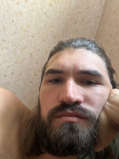 A close-up, low-angle, selfie-style shot features a man with long, dark hair, a full beard, and a mustache. He is resting his head on his right hand, which is propped against his cheek. His left arm is visible, bent and resting behind his head. The man's eyes are open and directed towards the camera. The background is a textured, light-colored wall, possibly wallpapered. The image appears to have been taken indoors. No specific location in Chernobylets, Russia, can be identified from the visual information provided.