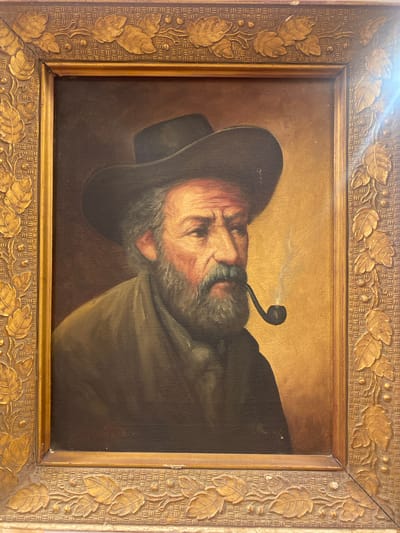 This is a framed oil painting of a man wearing a hat and smoking a pipe. The man has a beard and is looking to the right. The painting is housed in an ornate gold frame with leaf motifs. The background of the painting is a gradient of brown and gold. There is a faint plume of smoke rising from the pipe. The painting is displayed in an indoor setting. There is no indication in the image that this painting is located in the city of Legenda, Egypt.