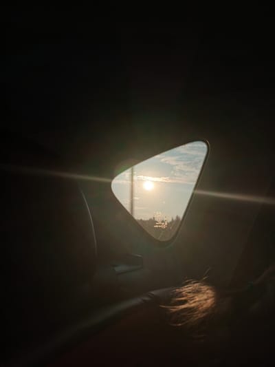 The image is a close-up, low-angle shot taken from inside a vehicle, likely a car, in Teplyaki, Russia. The primary focus is a triangular window, through which a bright sun is visible in the sky. The sun is low in the sky, suggesting either sunrise or sunset, and is casting lens flare across the window and into the interior of the car. The sky outside the window contains scattered clouds. Below the sky, a line of trees is visible. The window itself has water droplets on it and some markings, including a Mazda logo and other text, indicating it's likely a car window.
The interior of the car is mostly dark, with the exception of the light from the window. In the foreground, to the right, is the back of a person's head, with long, light-colored hair illuminated by the sunlight. The hair appears windswept. A portion of a dark, leather car seat is visible in the lower left of the frame. The overall impression is one of a journey, with the sun's glow creating a warm, atmospheric effect within the dim interior of the vehicle. There is no overt indication of any activity other than the passive observation of the external environment through the window.