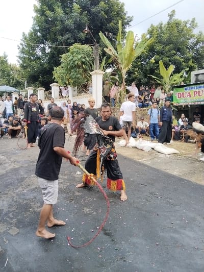 The image displays an outdoor cultural performance or ritual occurring on a dark, wet asphalt surface in Sumberejo, Indonesia, under an overcast sky. Two barefoot adult males are positioned centrally. The male on the left wears a black t-shirt and light-colored shorts, holding a braided red whip with a yellow handle extended towards the other participant. The male on the right faces him, wearing a black t-shirt, dark pants with red and yellow fringed cuffs, and an elaborate dark mask or headpiece resembling an animal's head, which he holds with both hands at chest level. He is in a wide, bent-knee stance. Red and pink petals are scattered on the ground around the two individuals.

A large, diverse crowd of onlookers, consisting of adults and children, observes the event from the background and midground. On the left, spectators stand behind a white decorative wall topped with a golden fence. On the right, individuals are seated on a raised dirt embankment, near a partially visible green banner for a "WARUNG SEMBAKO" (grocery stall) and "DUA PU". In the background, dense green foliage and several banana trees are visible. A street light fixture is mounted on a pole above the white wall. Several white sacks are present on the ground to the right of the central performers. Other individuals, some in dark attire and holding similar whips, are visible among the crowd, suggesting their participation in the event.