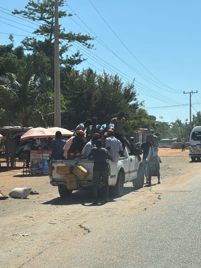 The image shows a densely packed group of people riding in the back of a white pickup truck. The truck is traveling on a paved road, and there are pedestrians walking alongside it. The truck bed also contains several yellow jerrycans. To the side of the road is a commercial stand selling PAGA branded products. The background includes trees and power lines against a clear blue sky. The location is likely Muxara, Mozambique.