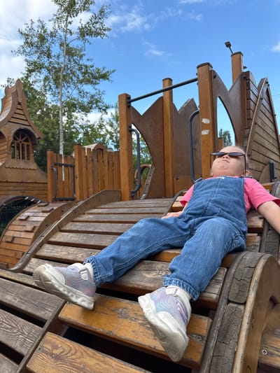 In this vertically oriented medium shot, a young child is lying down on a wooden playground structure. The child, wearing blue denim overalls over a pink long-sleeved shirt, has their legs extended and is looking upwards, away from the camera. They are wearing dark sunglasses. The playground appears to be made of weathered wood and has various levels and wooden planks. In the background, the sky is a clear blue with some scattered white clouds. Green trees with sparse foliage are visible, suggesting a natural outdoor setting. The overall lighting and cloud cover indicate it is daytime, likely a sunny day. The child's relaxed pose and upward gaze might suggest a moment of rest or observation. The image seems to be taken in a park or a playground area. There is no visible text in the image. The location context provided is Верхнерусское, Russia.