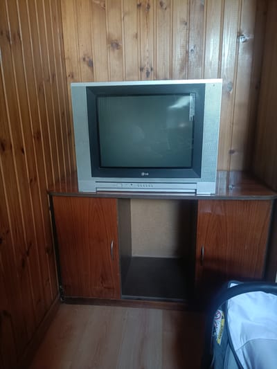 This is a full-shot, eye-level, interior photograph of a television on a wooden cabinet. The television is a CRT model with a silver frame and a gray screen displaying a static image or reflection. It has the LG logo on the front. Below the television is a wooden cabinet with two doors and an open compartment in the middle. The walls are paneled with vertical wooden planks, and the floor is covered with light-colored laminate flooring. In the lower right corner of the image, a portion of a white car seat with an airbag label is visible. The scene appears to be indoors in a room with wooden walls and flooring. The location is identified as Oxentea, Moldova.
