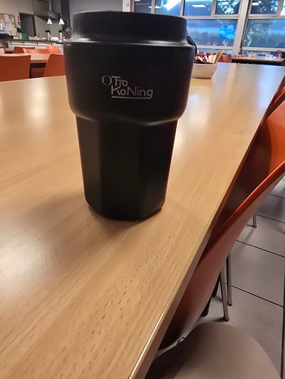 The image shows a black tumbler with the logo "OTRO Konin" on a wooden table. In the background, there are several orange chairs and a kitchen area with appliances. The location is a cafeteria or dining hall, likely in Lemberge, Belgium, given the context of the prompt. There are no people clearly visible in the foreground or mid-ground. The lighting suggests an indoor setting. The tumbler is the primary subject of the image, positioned in the center.