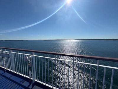The image captures a view from a ferry deck looking out at a body of water under a clear blue sky with the sun high overhead, creating a bright glare on the water's surface. The ferry's railing, made of white metal bars with a dark brown wooden top, runs along the bottom of the frame. A speaker and some electrical equipment are mounted on the railing. The blue deck of the ferry is visible in the foreground. In the distance, a long, tree-lined coastline is visible, dotted with what appear to be small islands. The sun's rays are visible as a lens flare, and a faint, curved white contrail stretches across the sky. The location is likely the waters surrounding Karlskrona, Sweden, given the presence of coastal islands and the ferry traffic common in this archipelago.