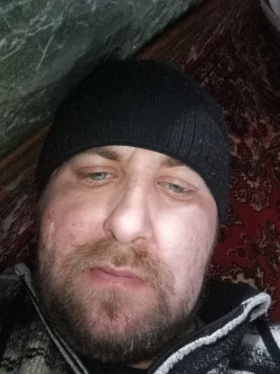 A close-up, slightly angled, color photograph captures a middle-aged male subject wearing a black beanie. The subject has light skin, a beard and mustache of mixed brown and gray hair, and light-colored eyes. His complexion displays some skin texture and minor blemishes. He's wearing what appears to be a jacket with a patterned exterior and a light-colored, possibly fleece, lining. The background shows a wall with a mottled green pattern, possibly wallpaper, on the left side. To the right of the subject's head is a patterned textile, possibly a carpet or rug, with a predominantly reddish-brown color and complex floral or geometric designs. No specific information available to confirm location in the city of Parabel, Russia.