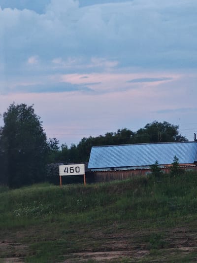 The image depicts a rural scene in Vorogovo, Russia, at dusk. The sky is a soft blend of blue and pink, suggesting either dawn or sunset. Fluffy clouds are scattered across the sky. In the foreground, a grassy slope leads up to a building with a corrugated metal roof, painted a light blue. The building appears to be made of wood. To the left of the building, a white signpost with the number "460" in black digits stands on wooden posts. Behind the building and signpost, a dense line of trees forms a dark backdrop, with a large, leafy tree to the far left. The overall impression is one of quietude and rural simplicity. There are no people visible in the image, and no specific activity is taking place. The lighting and colors suggest a peaceful evening in the countryside.