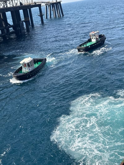 In Mers El Hadjadj, Algeria, two black tugboats navigate the deep blue ocean waters near a large, dark industrial structure. The structure, likely an oil rig or port facility, features multiple dark grey support beams and a walking platform above. The tugboats are engaged in operations, with one leaving a wake as it moves away from the structure, and the other closer to the viewer also creating a wake. The water is choppy, with white foam indicating the movement of the boats. The sky is clear and bright, suggesting a sunny day.