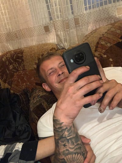 A man with a tattoo on his left arm and a visible injury on his right thumb is lying on a couch and taking a selfie with his phone. He is wearing a white t-shirt. The couch is covered with a patterned fabric. Behind the couch, a white curtain with a checkered pattern is visible. The location is Krushev Dol, Bulgaria.