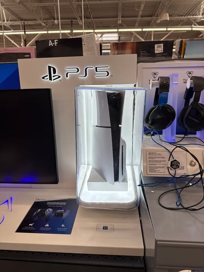 This media file captures a brightly lit retail display featuring gaming equipment, likely in an electronics or department store. The central subject is a white and black PlayStation 5 (PS5) console, displayed vertically within a clear, illuminated protective case. To the left of the console, a large, illuminated "PS5" logo with the PlayStation symbol is prominently featured. Below the PS5 console and logo is a dark, rectangular screen, likely a monitor or TV, possibly displaying game content or promotional material.

Above and to the right of the PS5 display, two pairs of black Turtle Beach headphones hang on a separate white display stand; one pair has blue accents. Entangled wires are visible near the headphones. Visible text on this display includes "TURTLE BEACH" and "RECON 200 GEN 2," indicating the specific model of headphones.

To the right of the PS5 display, a smaller poster or card promotes a "DUALSENSE EDGE" controller. A small "N" symbol, indicative of NFC (Near Field Communication), is also visible on a white surface nearby. In the background, typical retail shelving with various product boxes and overhead fluorescent lighting can be seen, along with a label that reads "A-F." The overall setting suggests a commercial environment designed to showcase gaming products.