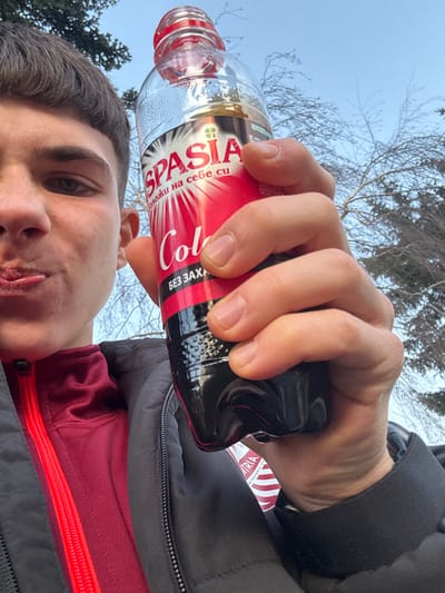 A young male is depicted in a close-up, low-angle view, holding a beverage bottle in what appears to be a selfie taken in Kolartsi, Bulgaria. The male, positioned on the left side of the frame and visible from the upper chest to just above his forehead, has short, dark brown hair. He exhibits a playful expression with his mouth partially open and tongue slightly protruding. He wears a dark, possibly black, zip-up jacket over a dark red long-sleeved garment with a bright red zipper. His right hand firmly grips a translucent plastic bottle containing a dark brown liquid, identified by its red and black label as "SPASIA Cola БЕЗ ЗАХАР" (sugar-free). The bottle's red cap is visible and appears slightly loose or partially open. The background consists of a clear, pale blue sky and numerous bare tree branches, suggesting a leafless season. A portion of a coniferous tree is visible on the far left.