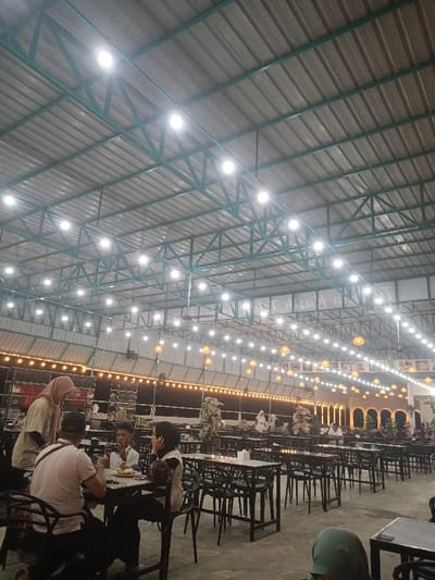 The scene depicts a large, high-ceilinged indoor or covered open-air dining establishment located in Matangjareung, Indonesia. The roof consists of corrugated metal supported by a prominent green metal truss framework. Numerous bright white LED lights are uniformly mounted to the trusses, providing primary illumination. Below these, additional decorative string lights with amber-toned, basket-weave lantern shades are strung horizontally across the space.

The floor is a dark, even surface. The dining area is furnished with numerous dark-colored rectangular tables and matching dark, slatted chairs, arranged in rows and clusters, indicating a substantial seating capacity. Hanging fans are visible attached to the ceiling trusses. In the background, there is a decorative architectural feature resembling a series of archways or windows with warm interior lighting.

Multiple patrons are present throughout the establishment. In the mid-ground to the left, four individuals are seated or standing around a table. A male in a white shirt is seated, facing the camera, holding a smartphone horizontally in both hands. Opposite him, a male in a light-colored shirt is seated with his back to the camera. To the right of the smartphone user, a female in a dark hijab and light-colored top is seated, facing left. A female in a light brown hijab and light-colored top stands to the far left of this group. Other patrons, including individuals wearing hijabs, are visible seated at various tables further in the background. A person wearing a dark green hijab is partially visible in the lower right foreground.