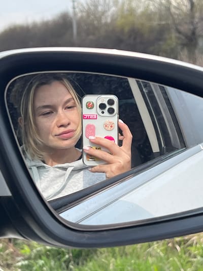 A blonde woman is captured in a selfie reflected in the side mirror of a car in Semeno-Aleksandrovka, Russia. Her face, seen from the chest up, is at the lower part of the reflection, angled slightly upwards, revealing a subtle smile and freckles. Her light blonde hair falls around her face.
Her left hand holds a smartphone, visible in the upper part of the mirror's reflection. The phone is encased in a clear cover and features several decorative elements on its back: a round popsocket with a floral pattern, a pink rectangular sticker, a yellow/gold rectangular sticker, and a red sticker displaying the white text "ERIC". Her fingernails are neatly manicured.
The mirror's reflection also shows parts of the car's interior, including a light-colored fabric of what appears to be a hoodie or sweatshirt worn by the woman, and the car's window frame. Outside the car, visible in the mirror and faintly in the background, is a blurred natural environment with green foliage and trees, suggesting a sunny daytime setting in an outdoor area.