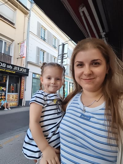 The image depicts an outdoor scene in front of a commercial establishment. A young girl with braided hair, wearing a black and white striped dress, stands to the left, smiling. To her right, a woman, likely her mother, also smiles at the camera. The woman is wearing a white and blue striped tank top and a silver necklace. In the background, a building with shuttered windows is visible, along with a sign for "Le Soleil de Boup" and images of food items. A person can be seen sitting on a low wall to the far left. The setting appears to be a street in Pomponne, France, likely a commercial district.