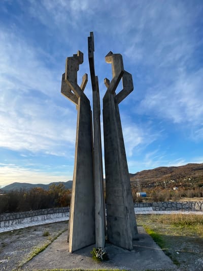The image depicts the Monument to the Fallen Fighters in Barutana, Montenegro, under a clear blue sky with scattered cirrus clouds. The central subject is a large, grey concrete monument composed of several tall, angular, brutalist-style pillars that converge and reach upwards, suggestive of stylized human figures. These concrete forms exhibit weathering. The monument stands on a concrete base, at the foot of which a small amount of green foliage, possibly a discarded wreath, is present.

The immediate foreground consists of a patchy grassy and dirt area surrounding the monument. A low, curved retaining wall constructed of stacked stones and concrete extends across the mid-ground, paralleled by a narrow concrete pathway. In the background, rolling hills covered with sparse, autumnal deciduous trees and some evergreen vegetation are visible. On the right side of the background, several distant white buildings and a tall metal pylon structure are discernible on a hillside. No individuals are present in the scene.