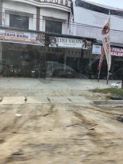 In Manetunong, Indonesia, a storefront displays multiple banners. One banner reads "BENGKEL MOTOR USAHA BARU," indicating a motorcycle repair shop. Another banner advertises "LYRA SALON," a hair salon. Further to the right, a banner for "HONDA PRIMA NUSANTARA" and "PENJUAL DEALER" suggests a Honda dealership. Several motorcycles are parked in front of the establishments. The scene is captured from a moving vehicle, resulting in motion blur. The environment appears to be a street in a commercial area, with a foreground of unpaved ground, possibly under construction or undergoing road work.