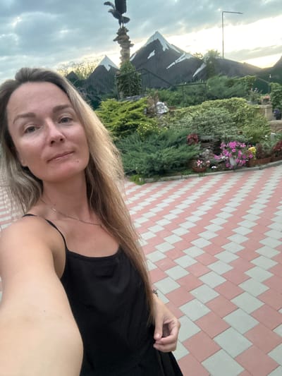 A woman with long blonde hair takes a selfie in front of a decorative garden scene. She is wearing a black tank top and a silver necklace. The background features artificial mountains, lush green bushes, and pink and white flowers in planters. A statue of an eagle perched on a rock column stands prominently in the upper left background. The ground is paved with a diamond pattern of pink and light gray tiles. The sky above is cloudy with hints of sunset light. The location is described as being in the city of Yaroslavskaya, Russia.