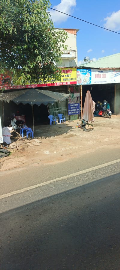 The image captures an outdoor scene in Ia Tôr, Vietnam, likely in front of a small business. A grey-blue tarped awning extends over a dirt and concrete area, providing shade for a workspace. Inside the shaded area, a red toolbox is visible on the left, and a white motorcycle is parked. Two bright blue plastic chairs are placed under the awning. In the foreground, a paved road runs diagonally across the lower part of the image, with a white line marking its edge.
Above the awning, a yellow sign with red lettering reads "PHÁT" and includes text in Vietnamese related to motorcycle repair and sales. Adjacent to this is another sign with an image of a drink bottle and Vietnamese text, possibly advertising beverages. Further to the right, a green building houses a storefront with signage that includes "XUÂN ĐÈN" and other Vietnamese text. A brown tarped object, possibly a piece of equipment covered for protection, stands in the open area. A red and white motorcycle is parked near the green building. A tall tree with green foliage dominates the upper left corner of the image. The sky is blue with scattered white clouds.