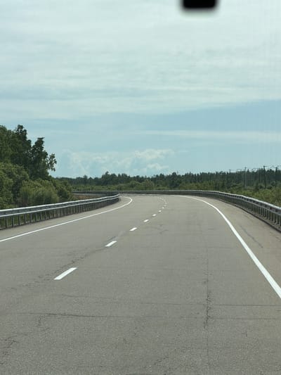 The image shows a curved asphalt road with white markings, bordered by a metal guardrail on the left and lush green trees on both sides. The sky is overcast with light grey clouds. The road appears to be a highway or a scenic route, with no vehicles or people visible. The surrounding environment is natural, with dense vegetation. The scene is captured from a driver's perspective, suggesting a moving vehicle. The location is identified as the city of Aur, Russia.